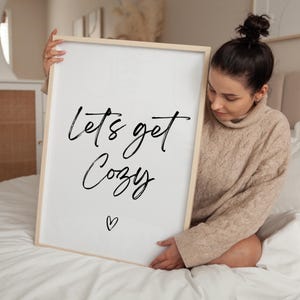 May include: A framed white print with the words "Lets get Cozy" in black script, with a small heart below. The frame is light wood. The print is held by a person wearing a beige sweater.