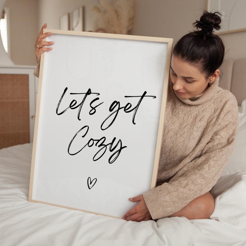 May include: A framed white print with the words "Lets get Cozy" in black script, with a small heart below. The frame is light wood. The print is held by a person wearing a beige sweater.