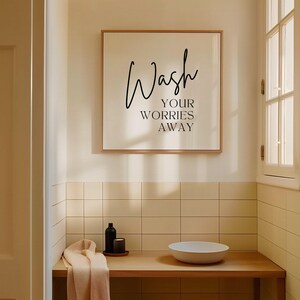 May include: A framed bathroom sign with the text "Wash your worries away" in black script font on a white background. The sign is hanging on a beige tiled wall above a wooden countertop with a white bowl sink and a bottle of liquid soap.