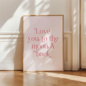Op de afbeelding: Een ingelijste kunstdruk met een lichtroze achtergrond en de tekst "Love you to the moon & back" in een zachtroze, decoratief lettertype. De lijst heeft een lichte houtkleur en de print leunt tegen een witte muur.