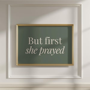 May include: A framed print with the text "But first she prayed" in a soft, elegant font. The text is in white against a muted green background, framed by a gold border. The print is mounted on a white wall with decorative molding.