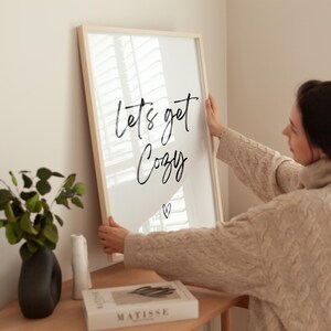 May include: A framed art print with the words "Let's get Cozy" in black script, with a small heart below. The frame is light wood, and the print is being held by a person wearing a beige sweater. Decorative items are on a wooden surface.