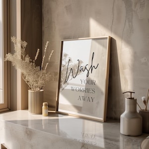 May include: A framed print with the text "Wash your worries away" in black script font on a beige background. The print is sitting on a light brown countertop with a small bottle and a soap dispenser.