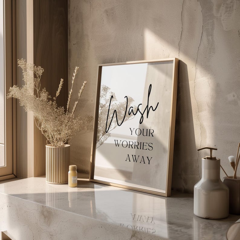 May include: A framed print with the text "Wash your worries away" in black script font on a beige background. The print is sitting on a light brown countertop with a small bottle and a soap dispenser.