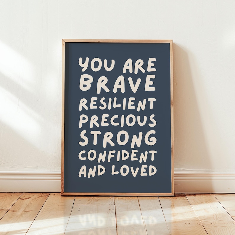 May include: A framed print with a navy blue background and white text. The text reads "YOU ARE BRAVE RESILIENT PRECIOUS STRONG CONFIDENT AND LOVED". The frame is a light brown color. The print is displayed on a wooden floor.