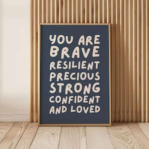 May include: A framed print with the words "YOU ARE BRAVE RESILIENT PRECIOUS STRONG CONFIDENT AND LOVED" in a white, handwritten-style font on a dark blue background. The frame is a light brown color, and the background is a wooden wall.