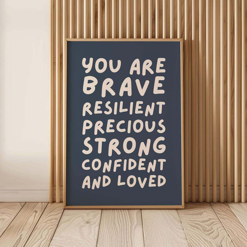 May include: A framed print with the words "YOU ARE BRAVE RESILIENT PRECIOUS STRONG CONFIDENT AND LOVED" in a white, handwritten-style font on a dark blue background. The frame is a light brown color, and the background is a wooden wall.