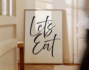 May include: A white framed poster with black calligraphy that reads "Let's Eat". The poster is hanging on a wall in a room with white trim and wood floors.