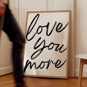 Puede incluir: Una impresión enmarcada con las palabras "love you more" escritas en cursiva negra sobre un fondo blanco.