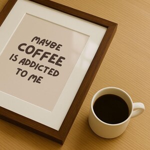 Könnte beinhalten: Ein gerahmter Druck mit dem Text "MAYBE COFFEE IS ADDICTED TO ME" liegt neben einer weißen Tasse mit dunklem Kaffee. Der Rahmen ist dunkelbraun und der Hintergrund hellbraun.
