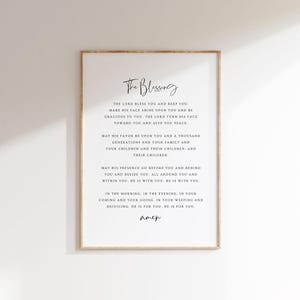 Può includere: Stampa incorniciata con il testo "The Blessing" in elegante scrittura. La stampa presenta una preghiera, con la parola "Amen" in basso. La cornice è di colore legno chiaro e lo sfondo è bianco.
