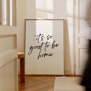 Puede incluir: Una impresión en lienzo enmarcada en blanco con caligrafía negra que dice "it's so good to be home". La impresión está colgada en una pared en una habitación con paredes blancas y pisos de madera.