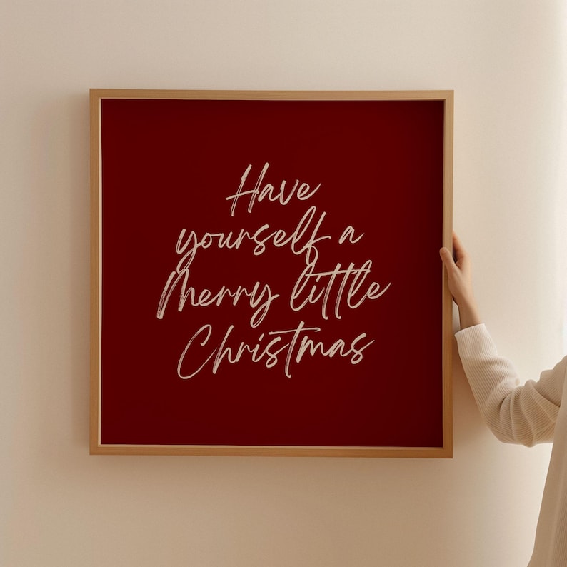 May include: A framed Christmas sign with a deep red background and the phrase "Have yourself a merry little Christmas" in white cursive script. The frame is a light wood color, and the sign is held by a person.
