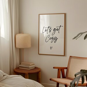 May include: A framed print with the words "Let's get Cozy" in black script, with a small heart below. The print hangs on a neutral wall, alongside a woven lamp and a wooden chair. The room has a warm, inviting atmosphere.