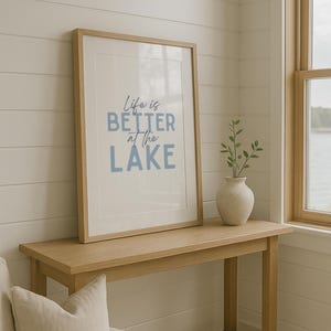 May include: A framed print with the text "Life is Better at the Lake" in blue lettering. The print is displayed on a light wood console table, next to a small vase with greenery. The room has white paneled walls and a window.