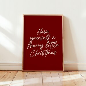 May include: A framed print with a deep red background and the white cursive text, "Have yourself a merry little Christmas." The frame is a light brown color. The print is displayed against a white wall and wooden floor.
