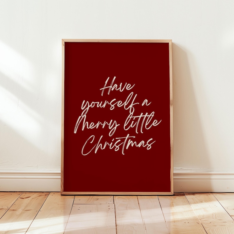 May include: A framed print with a deep red background and the white cursive text, "Have yourself a merry little Christmas." The frame is a light brown color. The print is displayed against a white wall and wooden floor.