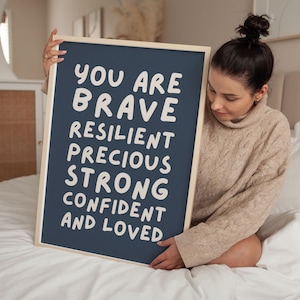 May include: A framed navy blue print with white text that reads "YOU ARE BRAVE RESILIENT PRECIOUS STRONG CONFIDENT AND LOVED." The print is held by a person wearing a beige sweater. The frame is a light wood color.
