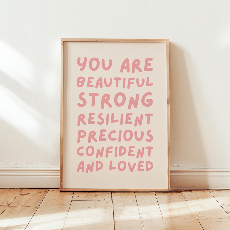 May include: A framed print with a white background and pink text that reads "You are beautiful strong resilient precious confident and loved".