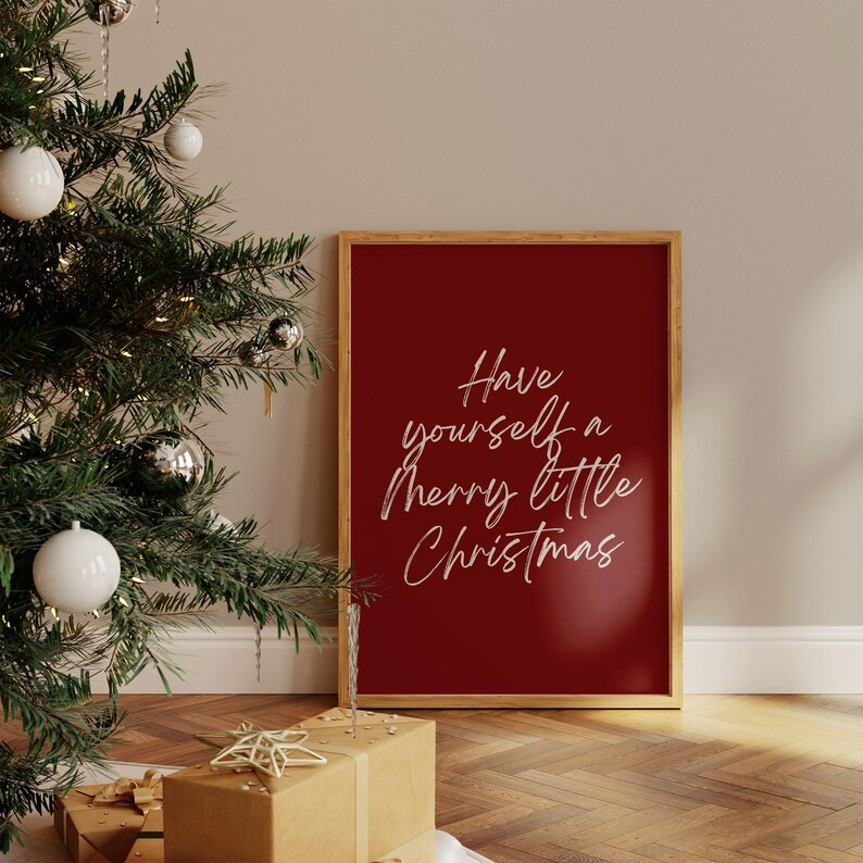 May include: A framed print with the text "Have yourself a Merry little Christmas" in white script on a deep red background. The frame is a light wood color. The print is displayed near a decorated Christmas tree and wrapped gifts.