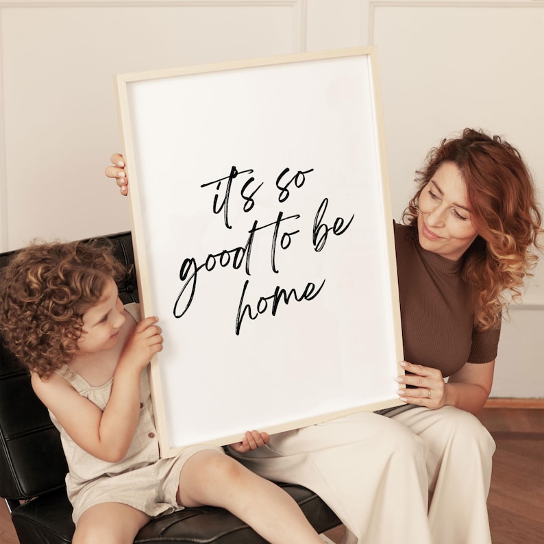 May include: A white framed print with black calligraphy that reads "it's so good to be home". A woman and a child are holding the print in front of a white wall.