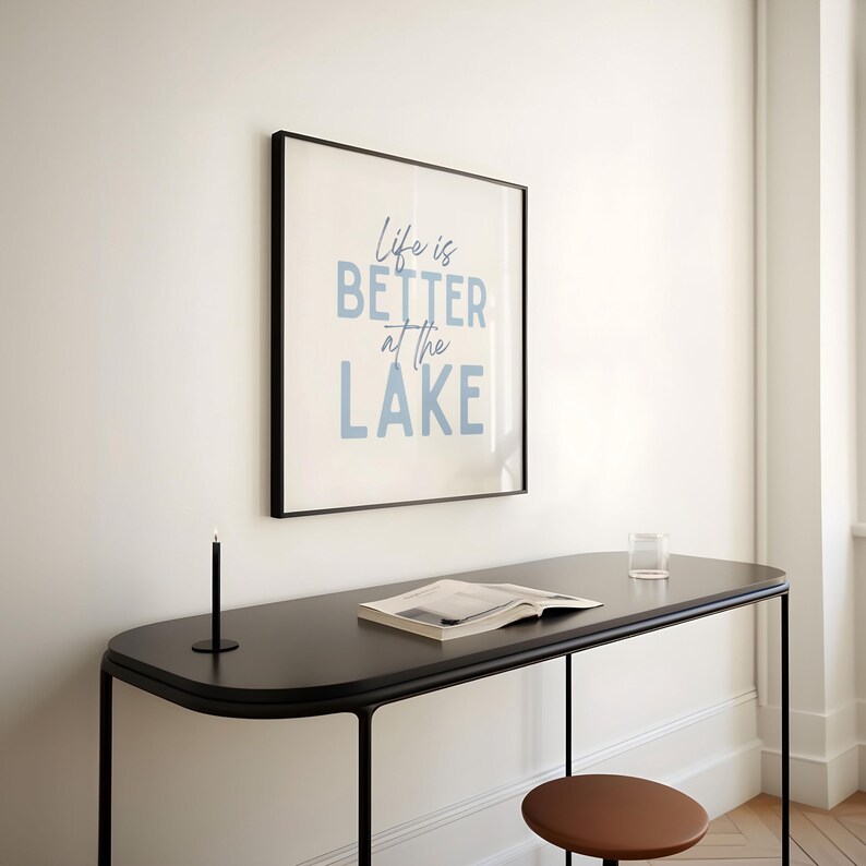 May include: A framed print with the text "Life is BETTER at the LAKE" in light blue lettering hangs on a white wall. Below, a black table with a book, glass, and candle holder. A brown stool is in the foreground.