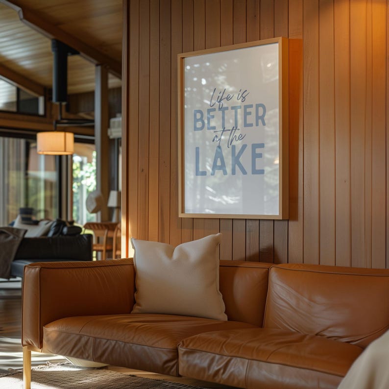 May include: A framed print with the text "Life is Better at the Lake" in blue lettering hangs on a wood-paneled wall. A brown leather sofa with a cream-colored pillow sits below the artwork, creating a cozy living space.