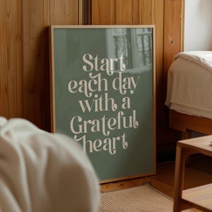 Puede incluir: Un póster enmarcado con el texto "Start each day with a Grateful heart" en una fuente blanca y elegante. El póster tiene un fondo verde salvia y un marco de madera. La imagen está en un dormitorio.