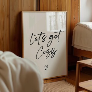 May include: A framed print with the words "lets get Cozy" in black script, with a small heart below. The print is in a light-colored wooden frame, set against a backdrop of wooden paneling and a bed.