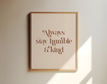 Puede incluir: Una impresión enmarcada con las palabras "Always stay humble & kind" en escritura marrón. La impresión tiene un fondo beige claro y un marco de madera. La imagen se muestra en una pared beige claro con una sombra.