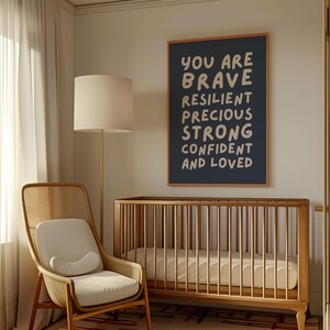 May include: A nursery scene featuring a wooden crib, a woven chair, and a floor lamp. A framed navy blue print on the wall reads "YOU ARE BRAVE RESILIENT PRECIOUS STRONG CONFIDENT AND LOVED" in white lettering. The room has a neutral color palette.