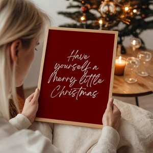 May include: A framed sign with the message "Have yourself a Merry little Christmas" in white script on a red background. The frame is a light wood color. The image is of a person holding the sign.