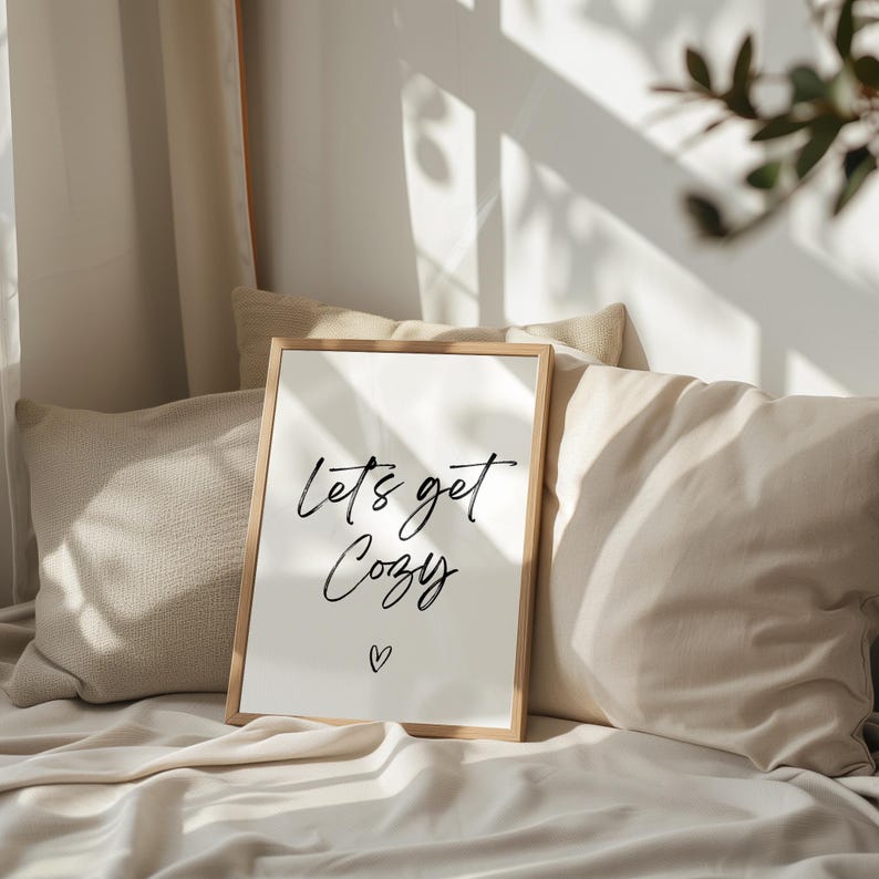 May include: A framed print with the words "Let's get Cozy" in black script, with a small heart below. The print has a light wood frame and is propped against pillows and bedding in neutral tones. The image evokes a sense of comfort and relaxation.