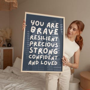 May include: A framed print with a navy blue background and white text that reads "YOU ARE BRAVE RESILIENT PRECIOUS STRONG CONFIDENT AND LOVED". The frame is a light wood color.