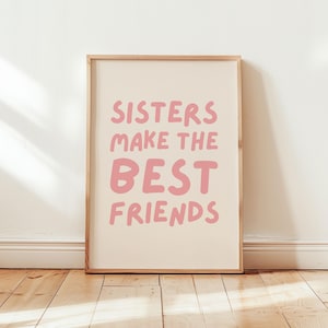 Może przedstawiać: Oprawiony wydruk z tekstem "SISTERS MAKE THE BEST FRIENDS" w różowych, bąbelkowych literach. Tło jest jasno-beżowe. Rama jest w kolorze jasnobrązowym. Wydruk jest wyświetlany na drewnianej podłodze.
