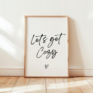 May include: A framed print with the words "Lets get Cozy" in black script, with a small heart below. The frame is a light wood color, and the background is white. The print is displayed against a white wall and wooden floor.