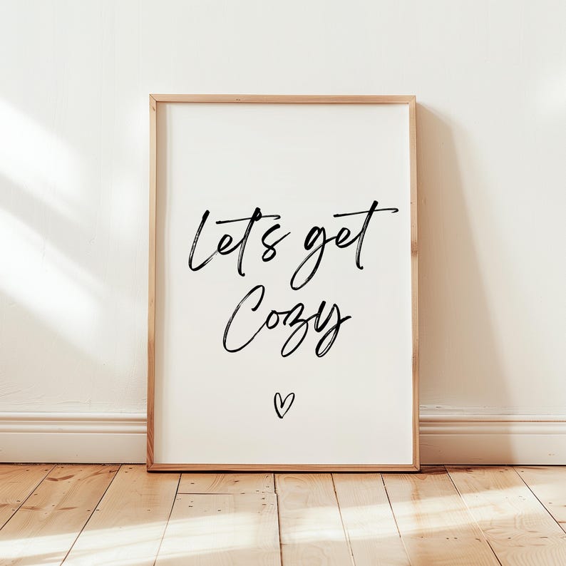 May include: A framed print with the words "Lets get Cozy" in black script, with a small heart below. The frame is a light wood color, and the background is white. The print is displayed against a white wall and wooden floor.