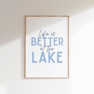 May include: Framed wall art with the phrase "Life is BETTER at the LAKE" in blue lettering. The artwork is in a light wood frame and hangs against a white wall. The text is in a mix of script and block letters.