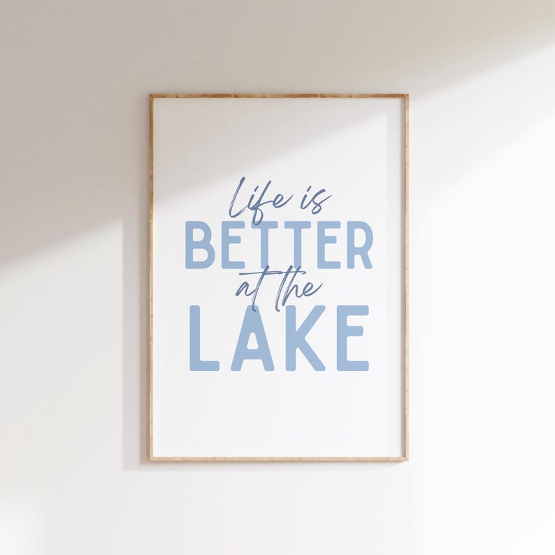 May include: Framed wall art with the phrase "Life is BETTER at the LAKE" in blue lettering. The artwork is in a light wood frame and hangs against a white wall. The text is in a mix of script and block letters.