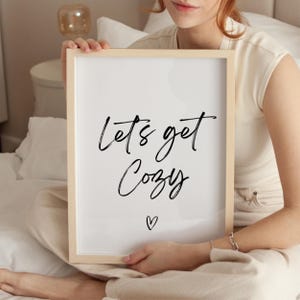 May include: A framed print with the words "Lets get Cozy" in black script, with a small heart below. The frame is light-colored wood. The print is held by a person wearing a cream-colored top.