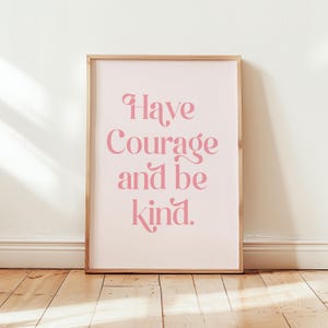 Puede incluir: Una impresión enmarcada con las palabras "Have Courage and be kind" en escritura rosa. La impresión tiene un fondo rosa claro y un marco de madera. La obra de arte se muestra contra una pared blanca y un suelo de madera.