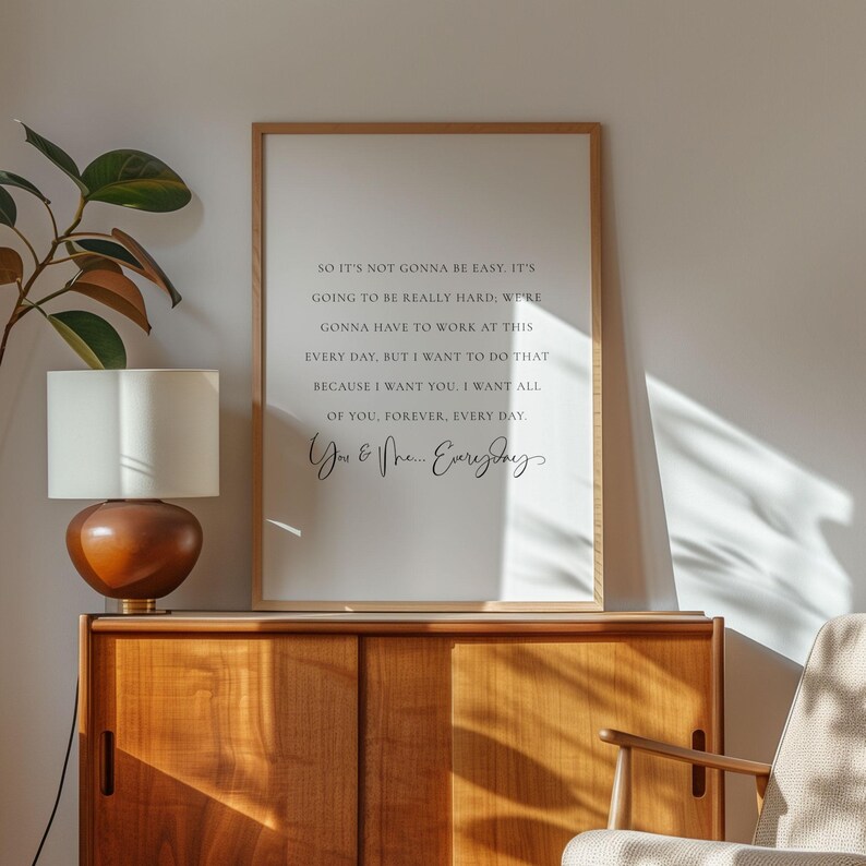 May include: A framed print with a light wood frame displays a romantic quote in black script on a white background. The text reads, "So it's not gonna be easy..." and ends with "You & Me... Everyday". The print is displayed on a wooden cabinet.