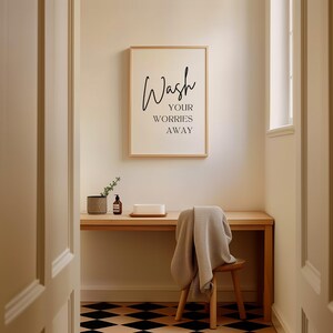 May include: A framed print with the text "Wash your worries away" hanging on a light beige wall in a bathroom. A wooden desk with a small plant, a bottle of liquid soap, and a white rectangular object is in the foreground. A wooden chair with a gray towel draped over it is in the foreground. The floor is black and white checkered tile.