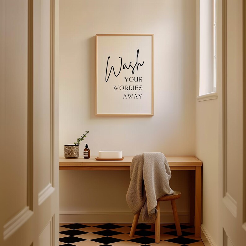 May include: A framed print with the text "Wash your worries away" hanging on a light beige wall in a bathroom. A wooden desk with a small plant, a bottle of liquid soap, and a white rectangular object is in the foreground. A wooden chair with a gray towel draped over it is in the foreground. The floor is black and white checkered tile.