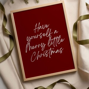 May include: A framed print with a deep red background and the text "Have yourself a merry little Christmas" in white script. The frame is a light wood color, and the print is surrounded by a cream-colored fabric and green ribbon.
