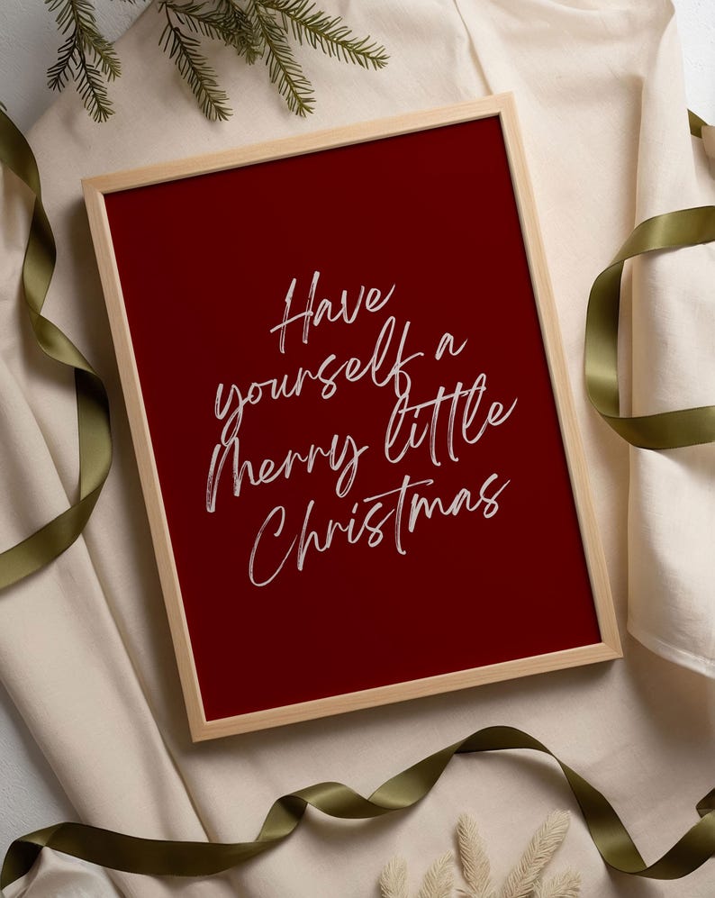 May include: A framed print with a deep red background and the text "Have yourself a merry little Christmas" in white script. The frame is a light wood color, and the print is surrounded by a cream-colored fabric and green ribbon.