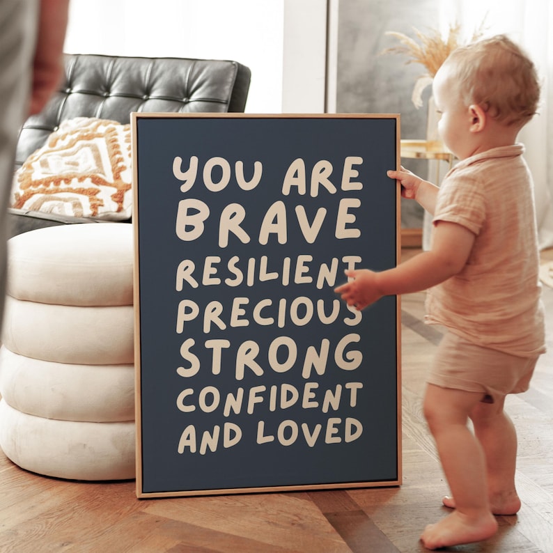 May include: A framed navy blue sign with the words "YOU ARE BRAVE RESILIENT PRECIOUS STRONG CONFIDENT AND LOVED" in white. A child is standing next to the sign. The frame is a light brown color.