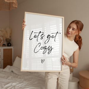 May include: A framed print with the words "Lets get Cozy" in black script, with a small heart below. The frame is light wood. The print is held up in front of a bed with white linens.