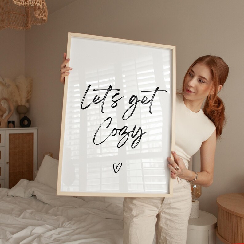 May include: A framed print with the words "Lets get Cozy" in black script, with a small heart below. The frame is light wood. The print is held up in front of a bed with white linens.