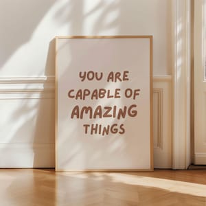 Puede incluir: Un cartel enmarcado con el texto "YOU ARE CAPABLE OF AMAZING THINGS" en letras marrones. El cartel tiene un fondo beige claro y un marco fino marrón claro. El cartel está apoyado contra una pared blanca.